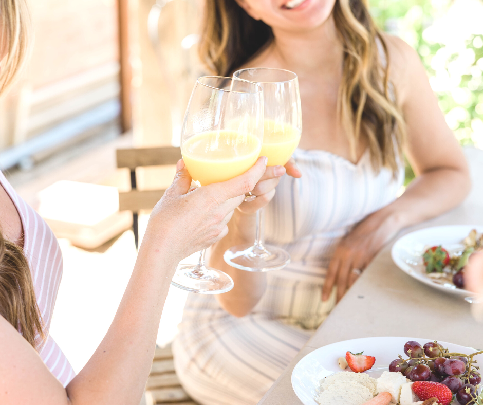 Two women cheers a glass of mimosa while enjoying a brunch on an outside patio. They have grapes, strawberries, and biscuits on the plate.