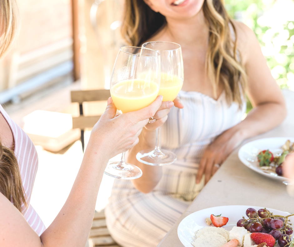 Two women cheers a glass of mimosa while enjoying a brunch on an outside patio.  They have grapes, strawberries, and biscuits on the plate.  